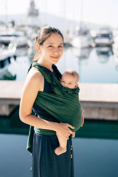 A smiling mother carries her baby in a baby carrier against a backdrop of a yacht.