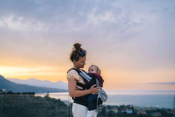 A mother carries her baby in a baby carrier at sunset.