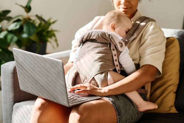 Woman working online on laptop while sitting on sofa with her baby carrier