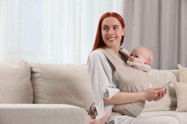 Mother with smartphone holding her child in sling (baby carrier) at home