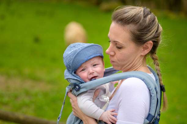 Happy woman looking to her crying baby while holding and carrying it in a baby carrier in a park