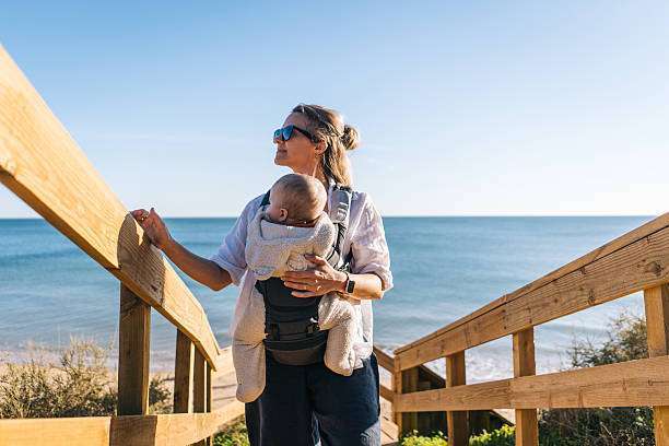 Mother holds baby son on hilltop above sea at sunset, baby carrier