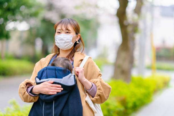 A mother is wearing a surgical mask and carrying her baby while on baby carrier